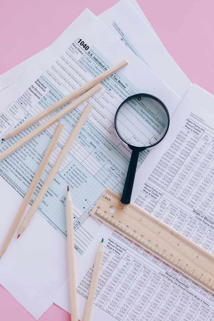 Tax documents with magnifying glass and pencils on a pink surface.